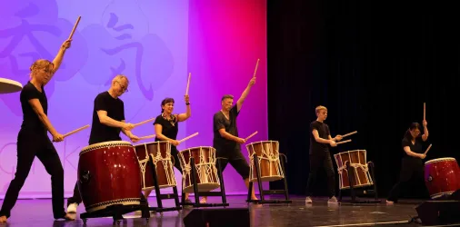 Eine Gruppe von Musikern spielt gemeinschaftlich auf großen Trommeln. Der Hintergrund ist in pinken und lila Tönen beleuchtet.