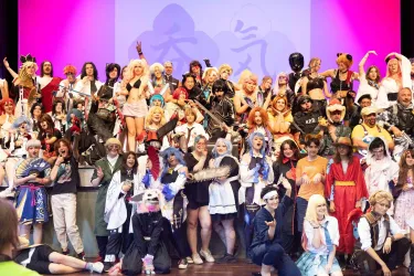 Eine große Gruppe von Menschen in bunten Kostümen steht auf der Bühne. Sie zeigen ihre Outfits und posieren fröhlich für das Foto.