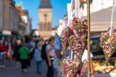 Hängende herzförmige Dekorationen in einer belebten Marktstraße. Im Hintergrund ist ein historischer Turm und viele Menschen zu sehen.
