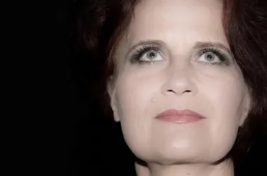 Eine Frau mit rotem Haar und intensiven Augen blickt nach oben. Ihr Gesicht ist sanft beleuchtet und wirkt geheimnisvoll.
