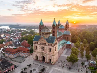 Eine beeindruckende Kirche mit Türmen steht vor einem malerischen Sonnenuntergang. Im Vordergrund sind charmante Häuser und Bäume zu sehen.