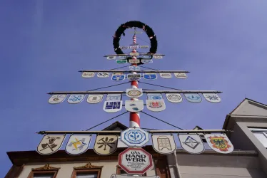 Ein bunter Maibaum mit verschiedenen Wappen und Symbolen steht vor einem Gebäude. Der Himmel ist klar und blau.