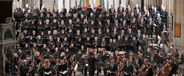 Ein großer Chor mit vielen Sängern steht in einer Kirche. Vor ihnen spielt ein Orchester auf der Bühne.