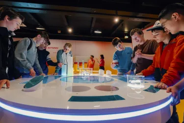 Eine Gruppe von Jugendlichen steht um einen interaktiven Tisch und experimentiert mit verschiedenen Objekten. Im Hintergrund sind weitere Personen und bunte Displays zu sehen.