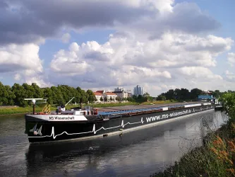 Ein Schiff mit dem Namen "MS Wissenschaft" fährt auf einem Fluss. Im Hintergrund sind Bäume und einige Gebäude zu sehen.