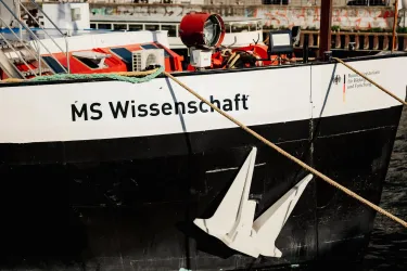 Ein Schiff mit dem Namen "MS Wissenschaft" ist am Hafen vertäut. Es hat eine schwarze und weiße Farbgebung mit einem auffälligen Anker.
