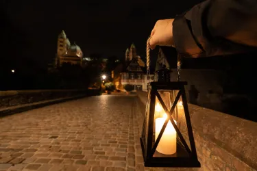Eine Person hält eine Laterne auf einem Steinweg bei Nacht. Im Hintergrund sind historische Gebäude und eine Burg zu sehen, die sanft beleuchtet sind.