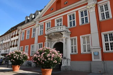 Ein schönes historisches Gebäude mit oranger Fassade und großen Fenstern. Vor dem Eingang stehen gepflegte, blühende Pflanzen in Töpfen.