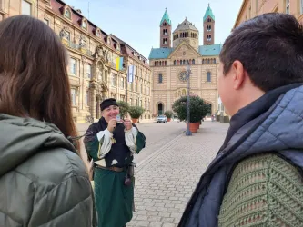 Ein junger Mann in historischer Kleidung spielt ein Instrument vor einem alten Gebäude. Im Vordergrund sind zwei Personen zu sehen, die ihm aufmerksam zuhören.
