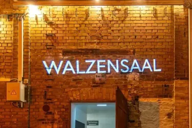Ein beschilderter Eingang zum Walzensaal mit leuchtendem Schriftzug. Die Wand ist aus hellen Ziegeln, und der Bereich ist gut beleuchtet.