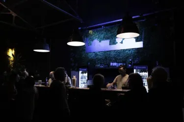 Eine stilvolle Bar mit einer beleuchteten Theke und Pflanzen an der Wand. Gäste stehen zusammen und unterhalten sich im schwach beleuchteten Raum.