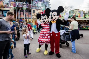 Zwei Personen im Kostüm von Mickey und Minnie Mouse posieren mit Kindern auf einem Fest. Im Hintergrund sind bunte Attraktionen zu sehen.