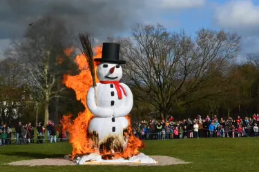 Ein brennender Schneemann steht mitten in einem Feld, umgeben von Zuschauern. Im Hintergrund sind Bäume und ein blauer Himmel zu sehen.