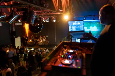 Eine lebendige Party in einem Club mit gutem Musik. Im Vordergrund steht ein DJ an den Turntables, während im Hintergrund Gäste tanzen und sich unterhalten.
