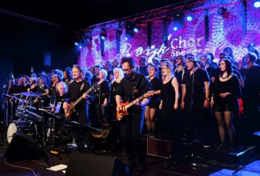 Eine lebendige Musikaufführung mit einer Band und einem Chor auf der Bühne. Die Musiker und Sänger tragen schwarze Kleidung und sind von bunten Lichtern umgeben.