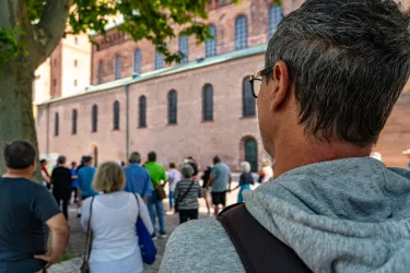 Eine Gruppe von Touristen steht vor einem historischen Gebäude. Im Vordergrund ist ein Mann zu sehen, der die Gruppe beobachtet.