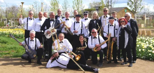 Eine große Gruppe von Musikern steht im Freien und posiert für ein Foto. Sie tragen schwarze und weiße Kleidung und halten verschiedene Instrumente in der Hand.