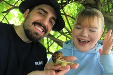 Ein Mann und eine Frau lächeln glücklich, während sie eine große Schnecke halten. Sie befinden sich unter einem grünen Baum.