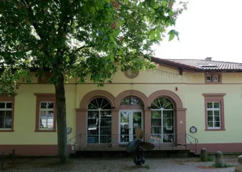 Ein historisches Gebäude mit großen Fenstern und einem gelben Anstrich. Im Vordergrund steht ein Baum, der Schatten spendet.