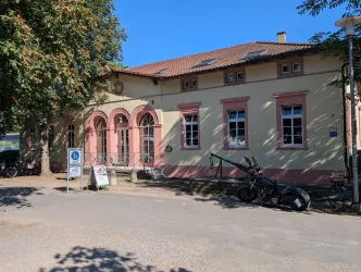 Ein historisches Gebäude mit einem hellen Außenanstrich in Gelb und Rot. Vor dem Gebäude stehen Informationsschilder und ein Fahrrad.