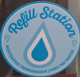 Ein rundes Schild mit der Aufschrift "Refill Station". Es informiert über kostenloses Trinkwasser mit einem Wassertropfen-Icon.