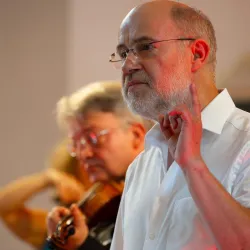 Ein Mann spricht und zeigt mit dem Finger nach oben. Im Hintergrund ist ein Mann mit einer Violine zu sehen.