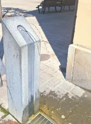 Ein Wassertrog aus Edelstahl steht an einer Straßenecke. Die Sonne scheint auf die Umgebung, und im Hintergrund sind Bänke zu sehen.