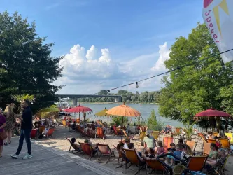 Eine entspannte Uferpromenade mit Liegestühlen und bunten Sonnenschirmen. Im Hintergrund fließt ein ruhiger Fluss unter einem blauen Himmel.