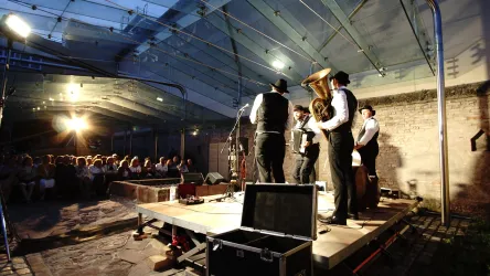 Ein Musikensemble spielt auf einer Bühne unter einem Glasdach. Das Publikum sitzt aufmerksam und genießt die Darbietung.