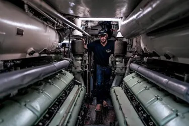 Ein Mann steht zwischen großen Motoren in einem Maschinenraum. Die engen Gänge und technischen Details zeigen die Komplexität des Innenlebens eines Schiffs.