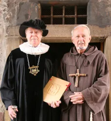 Zwei Männer stehen in historischen Kostümen. Der eine trägt eine schwarze Robe mit einem Kragen, der andere ist in ein braunes Mönchskostüm gekleidet.