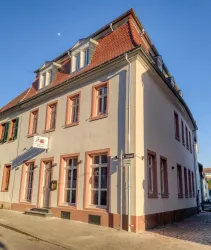 Ein historisches Gebäude an einer Straßenecke mit rot-braunen Fensterrahmen. Das Wetter ist klar und sonnig.
