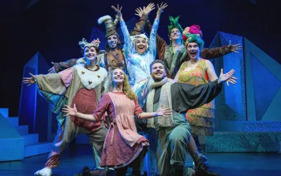 Eine fröhliche Theateraufführung mit mehreren Schauspielern in bunten Kostümen. Sie posieren gemeinsam auf der Bühne und strahlen Freude aus.