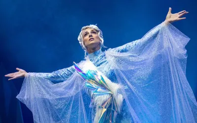 Eine beeindruckende Darstellerin in einem glitzernden, eisblauen Kostüm. Sie breitet ihre Arme aus und strahlt Selbstbewusstsein aus.