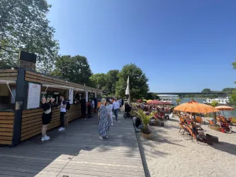 Ein belebter Strandbereich mit vielen Menschen und Liegestühlen. Sonnenschirme schützen vor der Sonne und im Hintergrund sind Bäume und Wasser zu sehen.