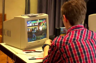 Ein junger Mann spielt ein Videospiel an einem älteren Computer. Auf dem Bildschirm ist eine actionreiche Szene zu sehen.