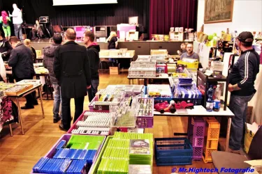 Ein Marktplatz mit mehreren Tischen, die mit verschiedenen Produkten wie Spielen und Spielzeug bedeckt sind. Menschen bewegen sich zwischen den Tischen und schauen sich die Angebote an.