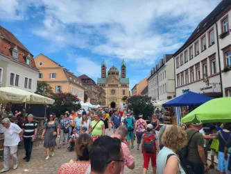 Eine lebendige Straßenszene mit vielen Menschen und bunten Marktständen. Im Hintergrund sieht man eine beeindruckende Kirche mit grünen Türmen.