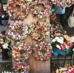 Bunte Blumengestecke in Form von Kränzen hängen an einem Holzbrett. Der Markt ist lebhaft mit weiteren Blumenarrangements umgeben.