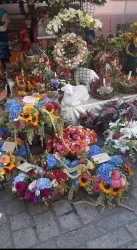 Ein bunter Markt mit verschiedenen Blumenarrangements und Kränzen. In der Mitte steht eine weiße Statue umgeben von Blumen.