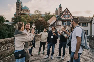 Eine Gruppe von Menschen feiert auf einer Brücke mit Gläsern in der Hand. Im Hintergrund sind historische Gebäude und Kirchen zu sehen.