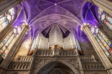 Eine eindrucksvolle Kirche mit hohen Gewölben und bunten Glasfenstern. Im Hintergrund ist eine große Orgel sichtbar, die von lila Licht hervorgehoben wird.