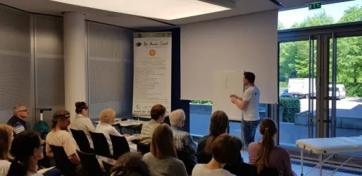Ein Mann präsentiert vor einem Publikum in einem Seminarraum. Im Hintergrund befindet sich ein Banner und große Fenster, die Tageslicht ins Zimmer lassen.