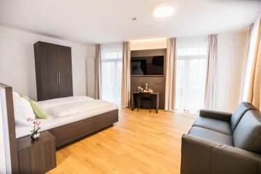 Ein modernes Schlafzimmer mit einem Doppelbett und einem grauen Sofa. Große Fenster lassen viel Licht herein und der Boden ist aus hellem Holz.