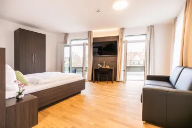 Ein modernes Zimmer mit einem Doppelbett und einem schwarzen Sofa. Große Fenster sorgen für viel Tageslicht und einen Blick nach draußen.