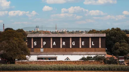 Ein großes Gebäude mit der Aufschrift "SPARGEL" ist vor einer ländlichen Kulisse zu sehen. Im Hintergrund erheben sich einige Bäume und ein blauer Himmel mit Wolken.