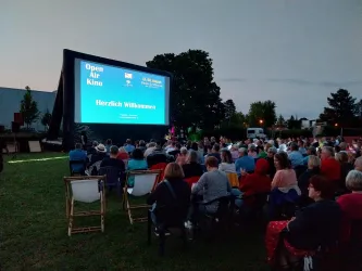 Eine Open-Air-Kinovorstellung mit vielen Zuschauern im Freien. Auf der Leinwand steht "Herzlich Willkommen" und es ist bereits dunkel.