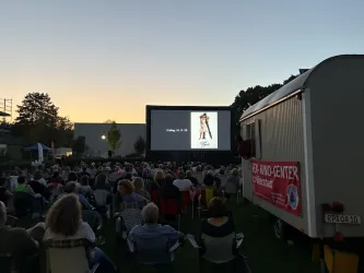 Eine große Menschenmenge sitzt im Freien vor einer Leinwand, auf der ein Film gezeigt wird. Im Hintergrund ist der Himmel bei Sonnenuntergang zu sehen.