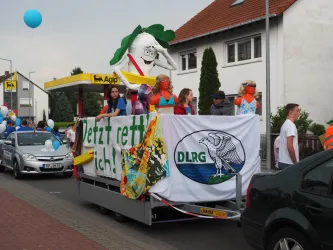 Eine bunte Parade mit einem geschmückten Fahrzeug und fröhlichen Menschen. Auf dem Fahrzeug ist ein Schild mit der Aufschrift "Jetzt rette ich!" und das DLRG-Logo zu sehen.