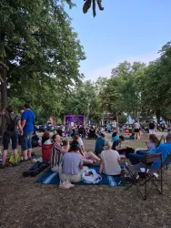Eine Menschenmenge sitzt und steht in einem Park und genießt eine Veranstaltung. Im Hintergrund ist eine Bühne mit bunten Lichtern zu sehen.